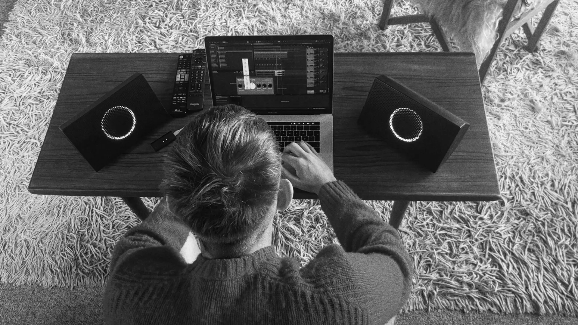 Vista cenital de una mesa con una persona en ordenador portátil MacBook Pro trabajando con un programa de audio. A cada lado del portátil hay una unidad de los monitores de estudio inalámbricos AIAIA UNIT-4 los primeros monitores profesionales inalámbricos del mundo. La foto está hecha en blanco y negro.
